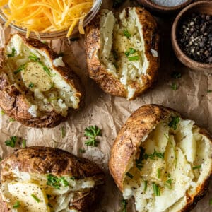 An overview shot of smoked baked potatoes filled with melted butter, chives, and parsley next to a bowl of shredded cheese.