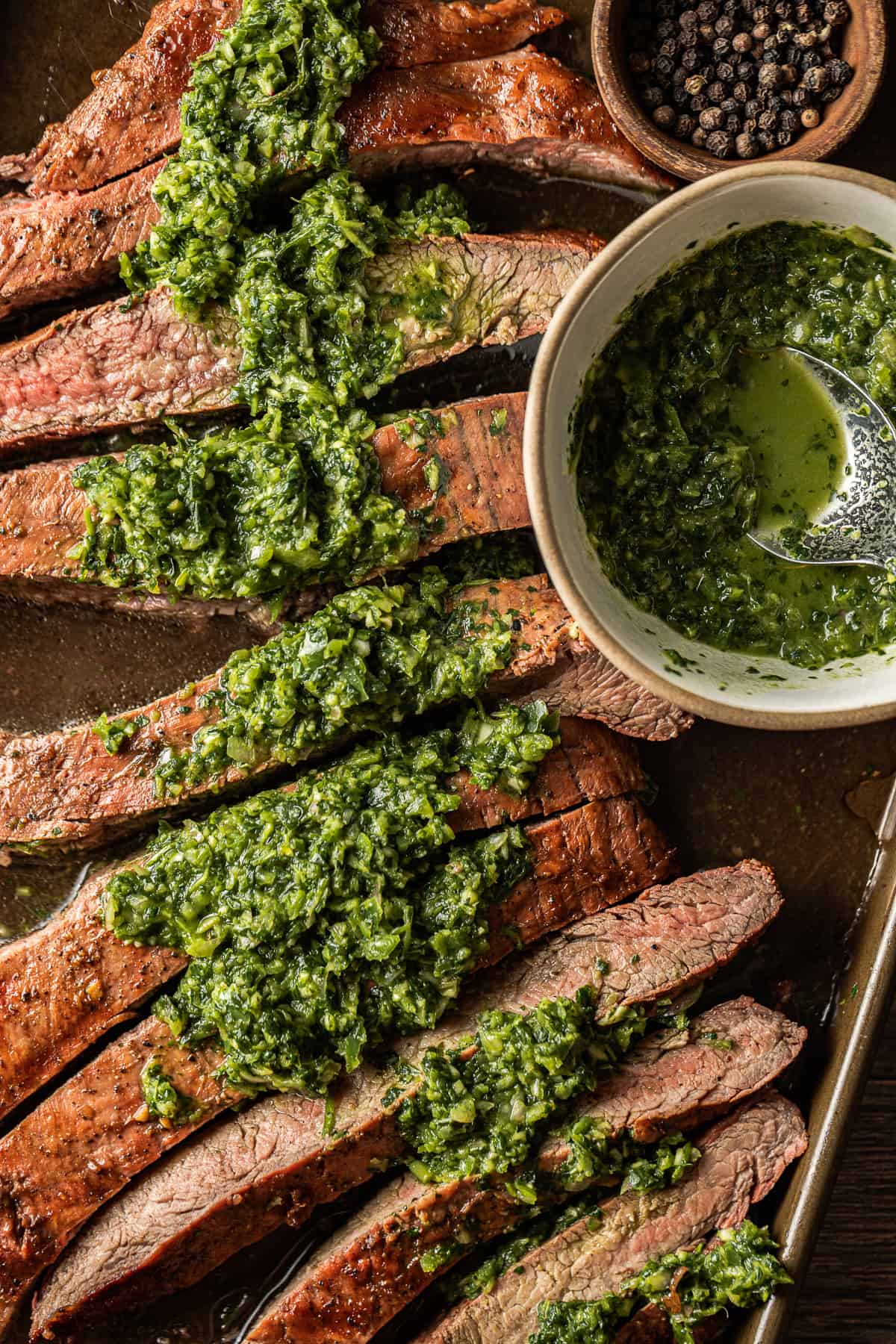 A sliced grilled flank steak topped with chimichurri next to a bowl of chimichurri near grilled avocado.
