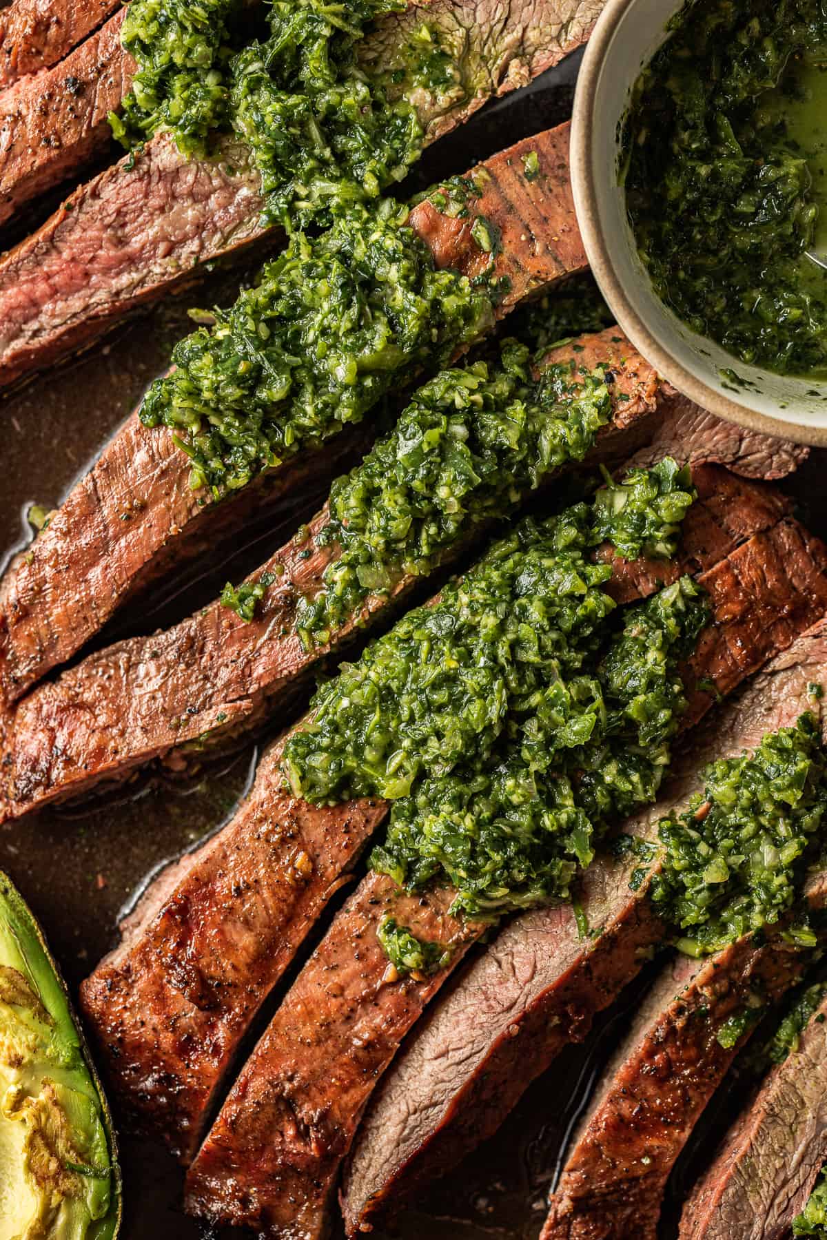 A sliced grilled flank steak topped with chimichurri next to a bowl of chimichurri.