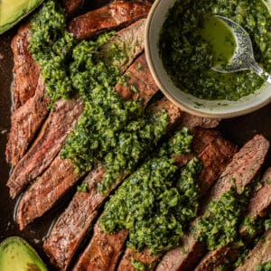 A sliced grilled flank steak topped with chimichurri next to a bowl of chimichurri.