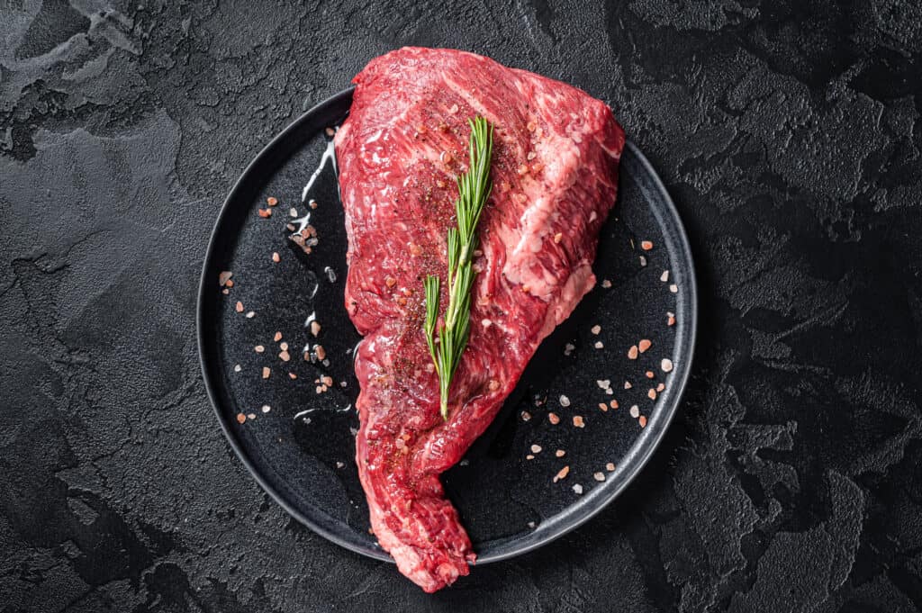 Seasoned raw tri-tip beef meat steak on plate. Black background. Top view.