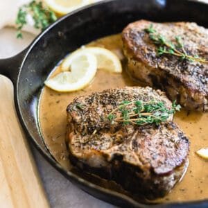 Two pork chops in a cast iron skillet with lemon garlic cream sauce, lemon slice, topped with thyme