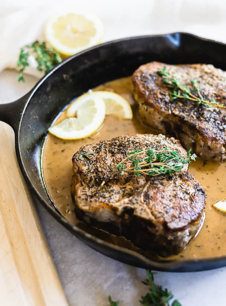 Two pork chops in a cast iron skillet with lemon garlic cream sauce, lemon slice, topped with thyme