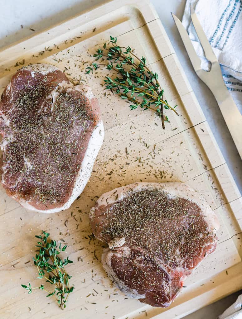 Two raw pork chops on a cutting board seasoned with salt, pepper, and thyme next to a linen and fork