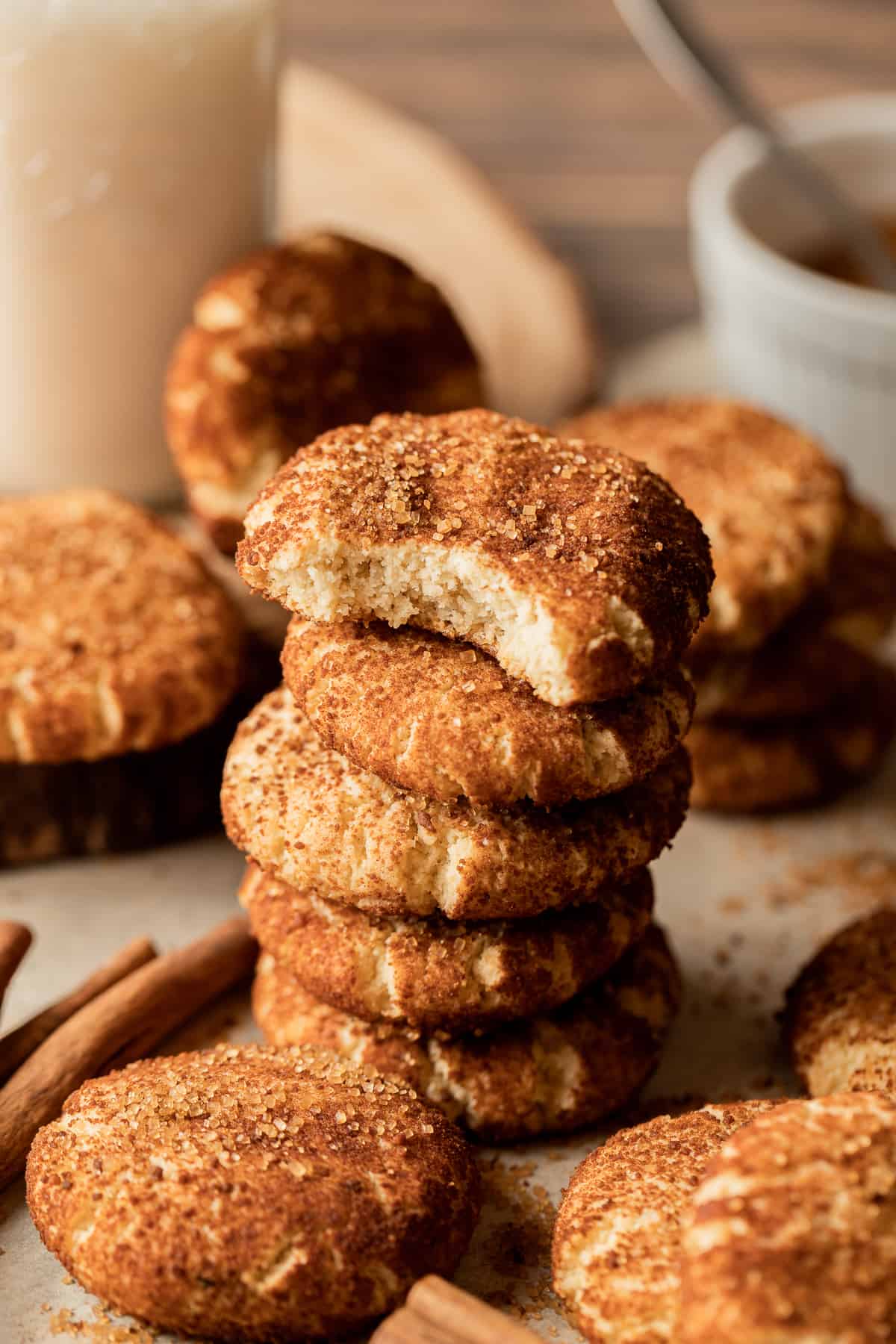 A stack of vegan snickerdoodle cookies with a bite taken out on a brown background near cinnamon sticks