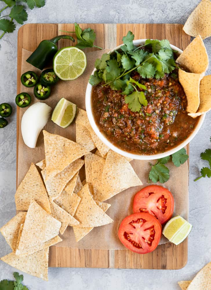 An overview shot of a bowl of salsa on a cutting board with salsa ingredients