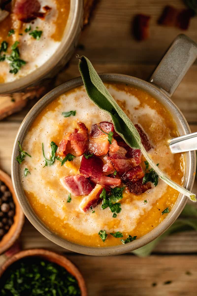 An overhead shot of creamy Whole30 butternut squash soup in a grey bowl on a wood background topped with coconut cream and bacon.