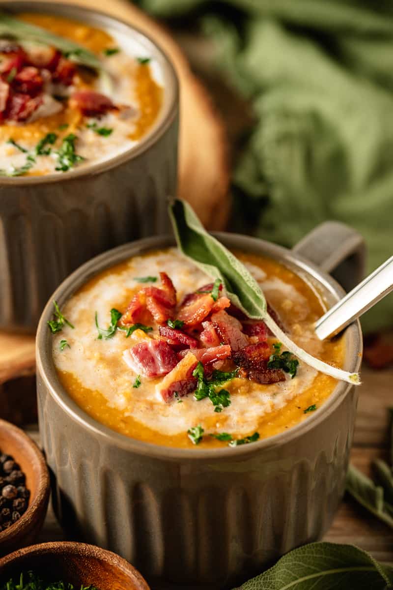 Two bowls of Whole30 Butternut Squash Soup topped with coconut cream, bacon, and fresh sage on a wood background