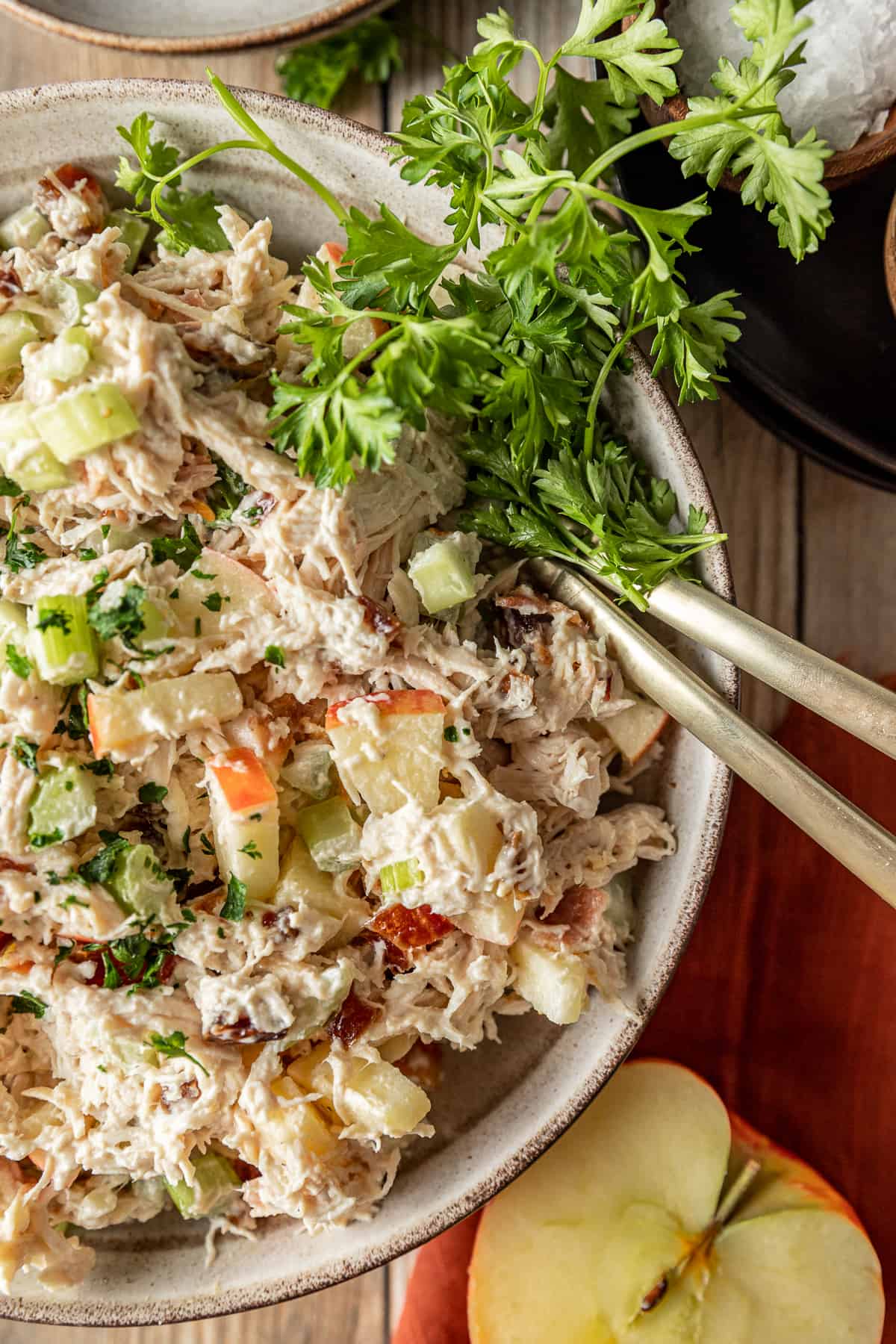 Chicken salad in a bowl on a wood background near chopped apples and topped with parsley.