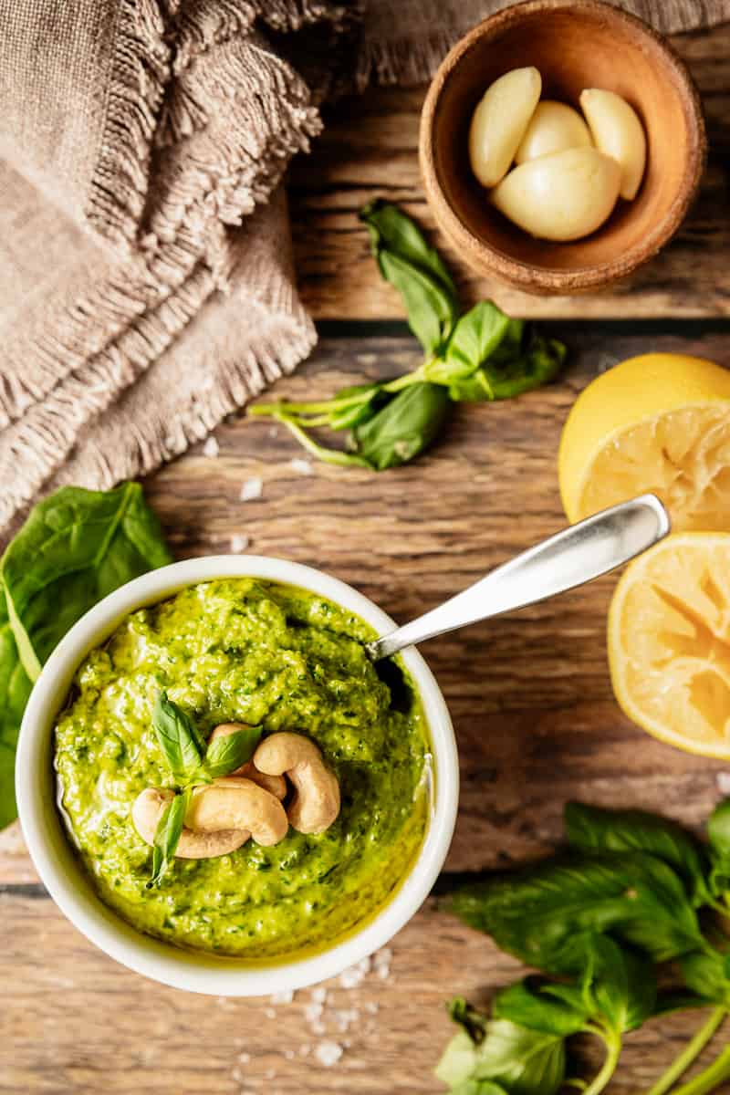 A bowl of whole30 pesto topped with cashews and olive oil on a wood background near basil, lemons, and garlic cloves
