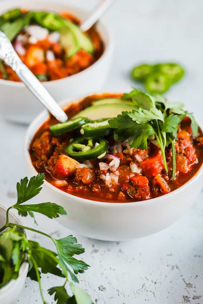 Two bowls of whole30 spicy sweet potato chili on a grey background next to a bowl of cilanto and topped with avocado and jalapeno