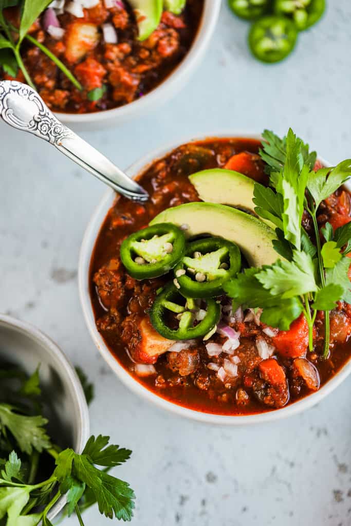 A top view of a bowl of whole30 spicy sweet potato chili on a grey background next to a bowl of cilanto and topped with avocado and jalapeno
