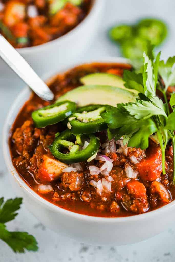 A bowl of whole30 spicy sweet potato chili on a grey background next to a bowl of cilanto and topped with avocado and jalapeno