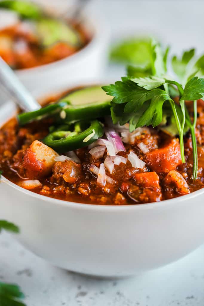 A close up of bowl of whole30 spicy sweet potato chili on a grey background topped with avocado and jalapeno
