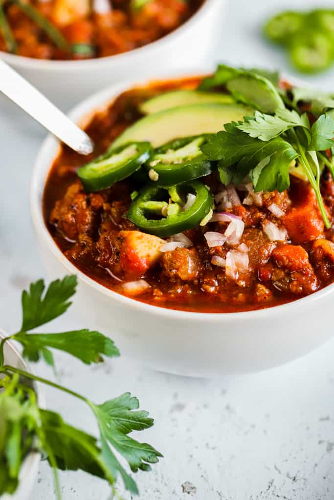A bowl of whole30 spicy sweet potato chili on a grey background next to a bowl of cilanto and topped with avocado and jalapeno