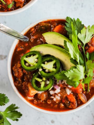 A bowl of whole30 spicy sweet potato chili on a grey background next to a bowl of cilanto and topped with avocado and jalapeno