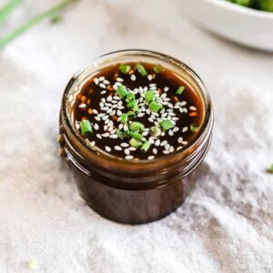 A jar of whole30 teriyaki sauce on a linen next to a bowl of broccoli