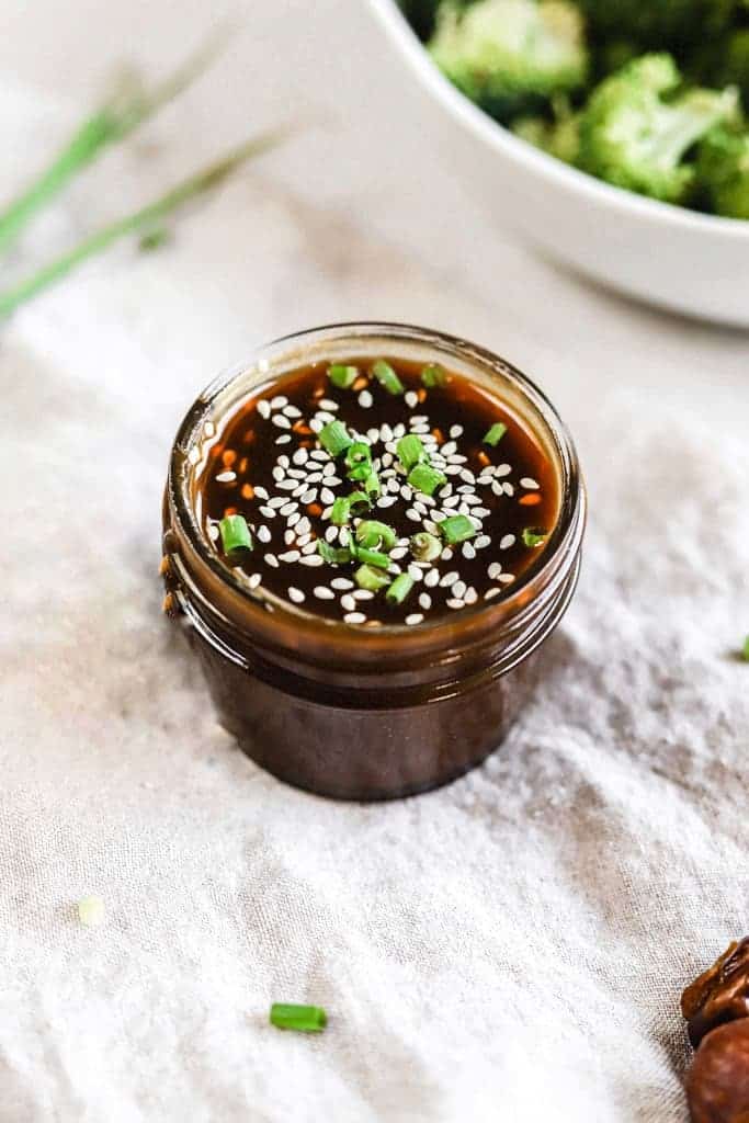 A jar of whole30 teriyaki sauce on a linen next to a bowl of broccoli