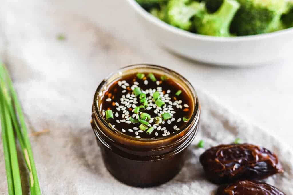 A jar of whole30 teriyaki sauce next to a bowl of broccoli and dates